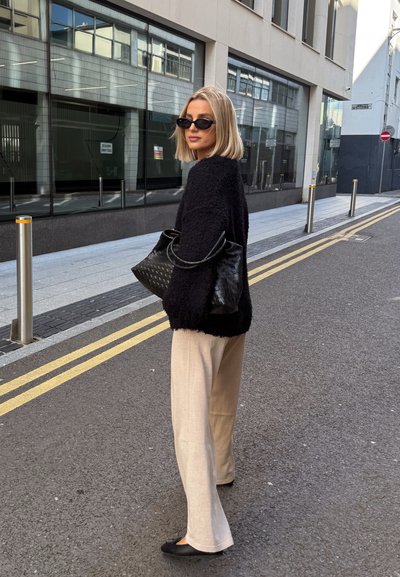 Suéter negro esponjoso, pantalones de pierna ancha beige, bolso negro y gafas de sol. Entorno urbano con edificios de cristal y pavimento visible.