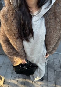 Felpa grigia e pantaloni della tuta sotto un soprabito di pelliccia sintetica marrone, con una borsa nera in vernice. Testure morbide e un look casual a strati.