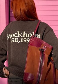 Άτομο με κόκκινα μαλλιά που φοράει καφέ πουλόβερ με το κείμενο "Stockholm SE, 1991", κρατώντας μια μεγάλη δερμάτινη τσάντα ώμου σε καφέ και βουργουνδί χρώμα.