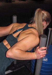 Atlētisks cilvēks tumšā sporta zālē, valkājot tirkīza krāsas sporta apģērbu. Veic dips vingrinājumu, izmantojot pelēkas stangas un zilu svara plāksni.