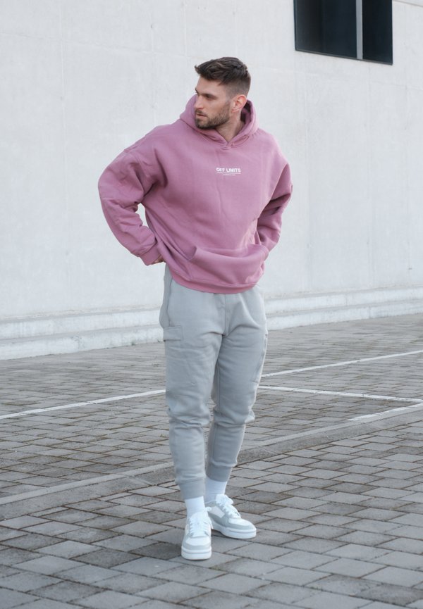 Hanorac roz cu textul "OFF LIMITS", asortat cu pantaloni jogger gri deschis și adidași albi. Materialul pare moale; design sportiv casual.