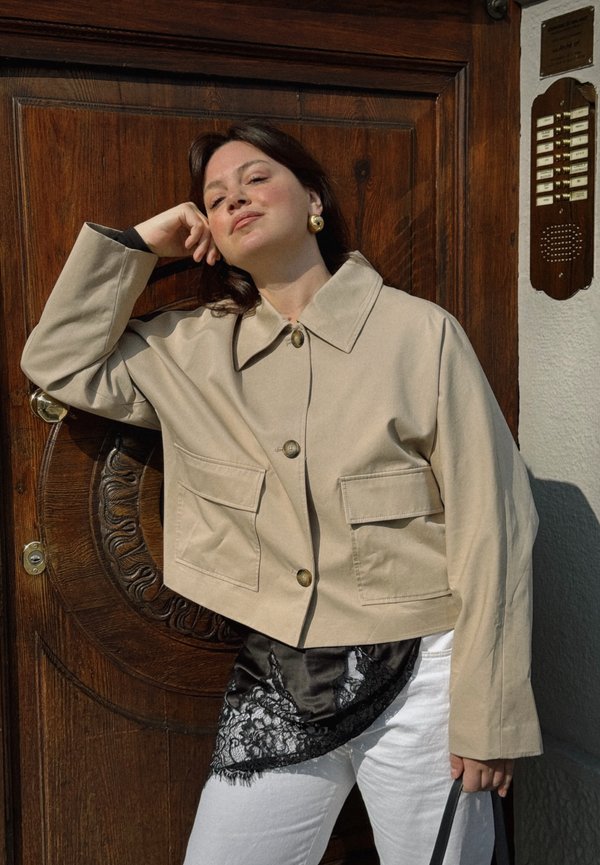 Donna appoggiata a una porta di legno, indossa una giacca beige, una maglietta in pizzo nera, pantaloni bianchi, orecchini dorati e tiene la tracolla di una borsa nera nella mano destra.