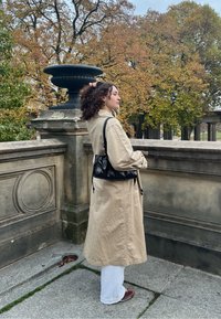 Μπεζ αμάνικο παλτό, μακρύς μανίκι, με χαλαρή εφαρμογή. Μαύρη τσάντα ώμου από δέρμα. Σγουρά μαλλιά, στηρίζεται σε μια πέτρινη αψίδα.