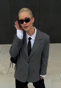 Grauer oversized Blazer, weißes Hemd, schwarze Krawatte, schwarze Sonnenbrille. Lange Nägel, silberne Ohrringe, hält eine kleine schwarze Handtasche. Einfarbiger Hintergrund.