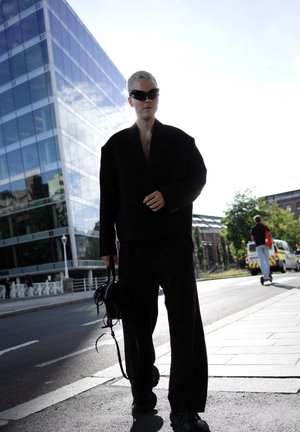 Blazer negro de gran tamaño con mangas anchas, pantalones a juego y gafas de sol negras angulares. Lleva un pequeño bolso negro. Entorno urbano.