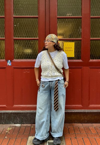 Jersey de color crema sobre una camiseta blanca, jeans de mezclilla anchos de color azul, con una corbata a rayas marrón y zapatos negros, de pie frente a una puerta roja.
