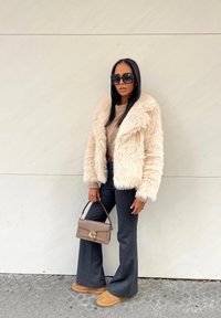 Chaqueta de piel sintética crema, suéter beige, pantalones de campana gris oscuro, botas marrones y un bolso marrón con detalles en dorado. Gafas de sol cuadradas.