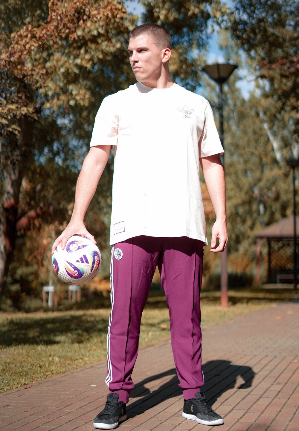 Tricou alb cu logo, pantaloni de trening maroon cu dungi laterale, pantofi sport negri, ținând o minge de fotbal colorată, cadru exterior.