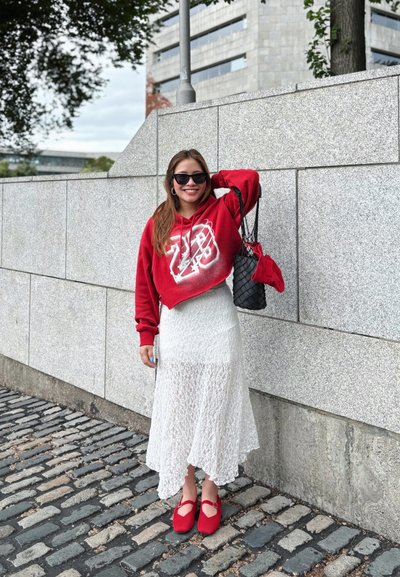 Sudadera roja cortada con gráfico blanco, falda maxi de encaje blanca, zapatos planos rojos, bolso negro, de pie contra una pared de piedra.