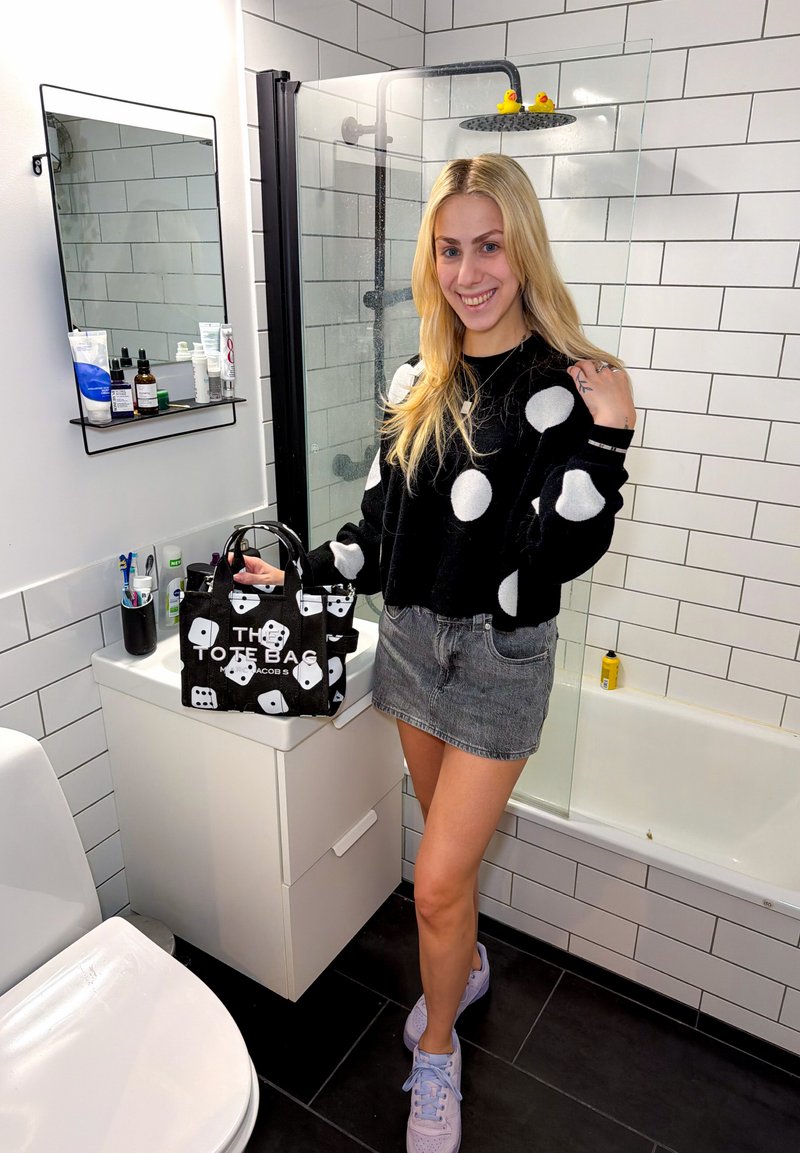 Γυναίκα με μαύρο πουλόβερ με πουά και γκρι φούστα χαμογελά κρατώντας τσάντα tote με μοτίβο ζαριών σε μαύρο και άσπρο σε μπάνιο με λευκά πλακάκια, ντουζιέρα και καθρέφτη.