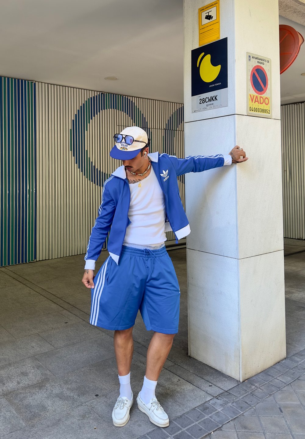 Giacca sportiva blu con dettagli bianchi e logo Adidas, abbinata a pantaloni corti blu coordinati. Una maglietta bianca e sneakers completano il look.