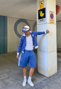 Giacca sportiva blu con dettagli bianchi e logo Adidas, abbinata a pantaloni corti blu coordinati. Una maglietta bianca e sneakers completano il look.
