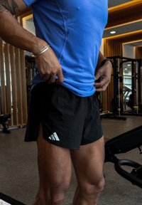 Sorte atletikshorts med sidesplitter og et reflekterende hvidt Adidas-logo, kombineret med en blå fugttransporterende T-shirt i en fitnesscenterindstilling.
