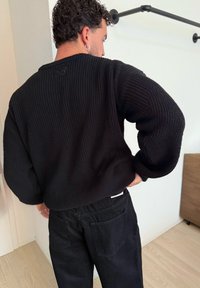 Suéter negro de punto con corte relajado, con un ribete texturizado. Combinado con pantalones negros, mostrando la vista trasera.