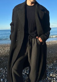Manteau noir texturé à larges revers, boutonné double rangée, associé à un pull noir côtelé et un pantalon gris foncé, sur une plage de galets.