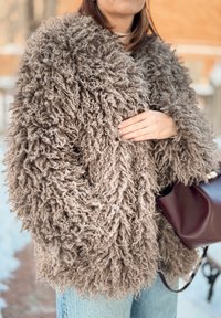 Veste duveteuse de couleur taupe, faite d'un tissu texturé et bouclé. Remarquable pour son apparence ébouriffée et sa coupe ample et décontractée. Portée avec un denim.