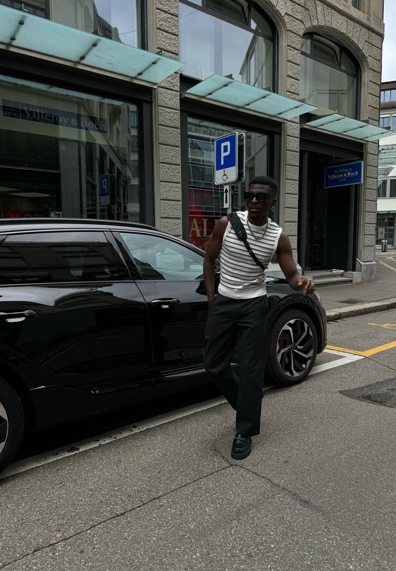 Μαύρο SUV παρκαρισμένο σε έναν δρόμο. Ένα άτομο με άσπρο ριγέ χωρίς μανίκια μπλουζάκι και μαύρα παντελόνια στέκεται δίπλα του, φορώντας γυαλιά ηλίου και τσάντα χιαστί.