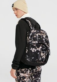 Rygsæk med sort bund, der har et sprøjte-mønster i beige og hvid. Inkluderer polstrede stropper og en frontlomme med lynlås.