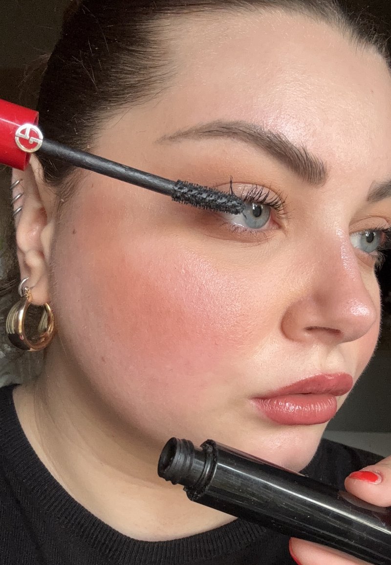 Tube de mascara noir tenu devant l'œil, avec un logo rouge. L'applicateur présente des poils épais, allongeant les cils. Teint de peau clair avec une légère touche de rose.