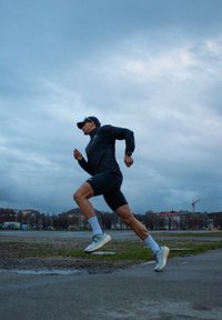 Vīriešu skrējējs melnā jakā un šortos, ar baltām zeķēm un gaišām sporta apaviem, skrien pa asfaltētu virsmu zem apmākušās debess.