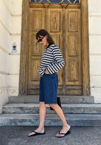 Mulher a usar uma camisa de manga longa às riscas, calções de ganga, chinelos pretos e óculos de sol, a passar por uma porta de madeira envelhecida com degraus de pedra.