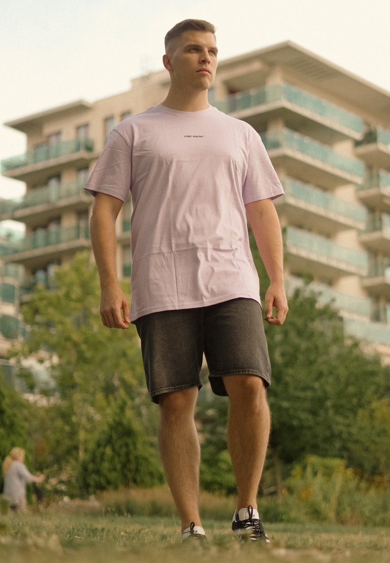 Jeune homme portant un t-shirt rose clair et un short foncé, debout à l'extérieur avec des immeubles d'appartements modernes et des arbres en arrière-plan.