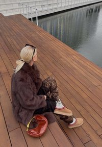 Smeđa umjetna krzna jakna, hlače s uzorkom leopard tiska, svijetlosmeđi platforme s crvenim detaljima i smeđa kožna torba na drvenim daskama uz vodu.