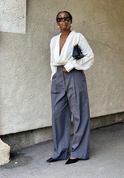 Mujer con blusa blanca drapeada, pantalones grises de talle alto y pierna ancha, zapatos negros puntiagudos, bolso negro, pendientes dorados y gafas de sol negras, de pie al aire libre.