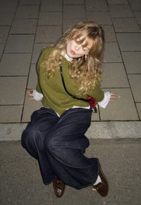Jeune femme aux longs cheveux bouclés assise sur le bord du trottoir, portant un pull vert, un large pantalon sombre et des chaussures marron, les yeux fermés et les lèvres pincées.