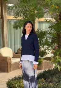 Femme aux cheveux foncés portant un pull bleu marine et une jupe noir et blanc, debout près de chaises en osier et d'un arbre vert à l'extérieur.