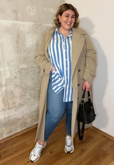 Abrigo trench beige sobre una camisa de rayas azul y blanca, combinado con jeans azules y zapatillas blancas. Bolso negro con asas cortas.
