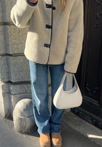 Casaco de fleece texturizado de cor creme com botões pretos, combinado com jeans azuis e sapatos slip-on castanhos. Segura uma pequena bolsa branca.