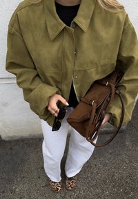 Masivna jakna od brušene kože maslinasto zelene boje, crni top, bijele hlače, smeđa torba i cipele s uzorkom leoparda. Dodaci uključuju crne sunčane naočale.