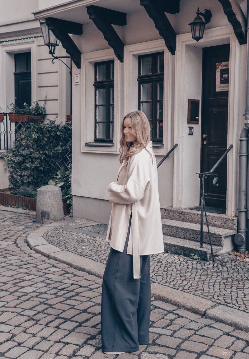 Manteau ample crème avec fentes latérales, porté sur un pantalon large gris foncé. Cheveux ondulés visibles sur fond de rue pavée et de bâtiment.