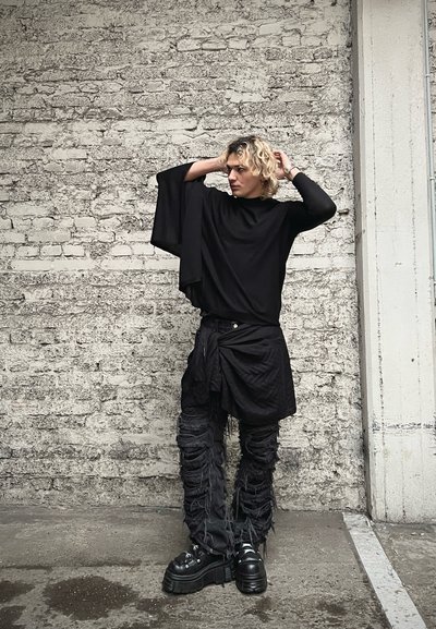 Blusa negra de gran tamaño con mangas amplias, combinada con pantalones negros texturizados que presentan detalles de flecos y zapatos de plataforma negros gruesos. Accesorios mínimos.