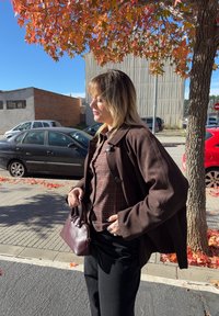 Hnedá semišová bunda cez károvanú košeľu, čierne džínsy a v ruke malá bordová kabelka. Na pozadí jesenné listy a autá.