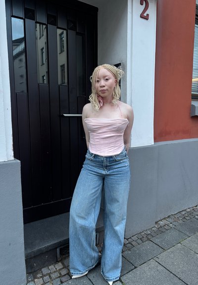 Top strapless de color rosa claro con detalles de fruncido; combinado con unos jeans de mezclilla de pierna ancha. De pie frente a una puerta de madera oscura.
