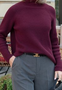Bordeaux gebreide trui met een hoge halslijn en ribbeltextuur, gecombineerd met grijze pantalons en een zwarte handtas. Gouden riemaccent.