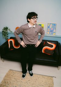 Brun rutig långärmad polo med vit krage, svarta byxor och svarta sneakers. Två mönstrade kuddar på en svart soffa i ett rum med mjuk textur.