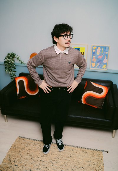 Polo de manga larga a cuadros marrones con un color blanco, pantalones negros y zapatillas negras. Dos cojines estampados sobre un sofá negro en una habitación de textura suave.