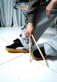 Czarne zamszowe sneakersy Puma z szarymi akcentami i gumowymi podeszwami, obok drewnianych pałeczek perkusyjnych trzymanych między nogami osoby w szarych spodniach.