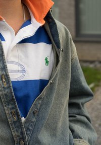 Casaco de ganga sobre uma polo riscada de azul e branco com um logótipo verde bordado. O colar é laranja, com botões de metal e variações de textura.