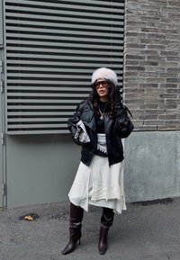 Schwarze Lederjacke über einem schwarzen Oberteil, weißer Rock mit Spitzenbesatz, dunkelbraune kniehohe Stiefel und ein flauschiger weißer Hut. Accessoires umfassen eine Halskette.