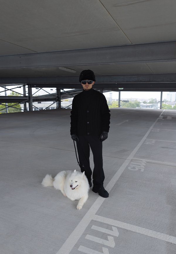 Un cardigan noir en tricot torsadé sur un col roulé noir, un pantalon noir et des lunettes de soleil. Un chien Samoyède blanc est assis à côté de lui sur le béton.