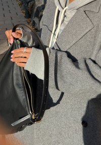 Palton gri din lână peste un hanorac gri, ținând o geantă din piele neagră cu fermoar și curea. Mâna arată un inel și unghiile vopsite în roz deschis.