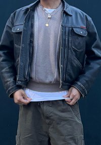 Fekete bőrkabát kontrasztos fehér varrással, világosbarna kötött pulóver fölött viselve, fehér inggel rétegezve, olívaké szatyor rövidnadrággal párosítva.