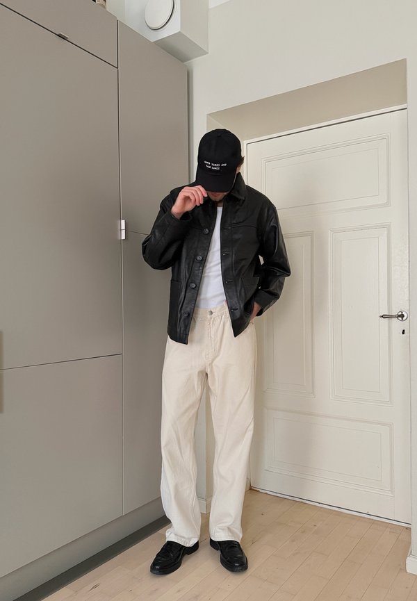 Persoon binnenstaand, gekleed in een zwart leren jas, witte broek, zwarte schoenen en een zwarte pet die het gezicht bedekt.