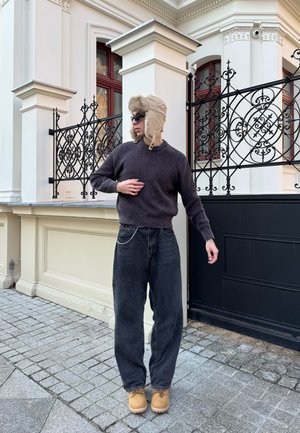 Gorro de piel de color beige, suéter oscuro de canalé, pantalones negros holgados con detalles de cadena plateada y botas de color marrón. Entorno urbano con una reja ornamentada de fondo.