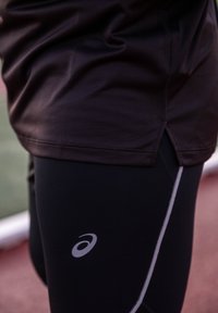 Person trägt schwarze Sportleggings mit weißem Asics-Logo und dunklem Sportshirt, steht auf einer Laufbahn.