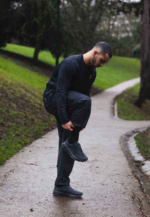 Hombre con ropa deportiva negra ajustándose la pernera del pantalón mientras está de pie en un camino sinuoso del parque, con césped y árboles cerca.
